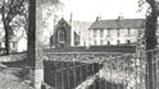 A woman in Edwardian dress stands next to a small set of mill buildings featuring a tall chimney stack. Behind is a row of dwellings and a church