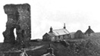 Black and white view of a rocky peninsula on which stand a group of single storey cottages arranged around the ruin of Old Slains Castle.