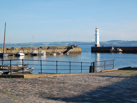 Colour view, taken on a sunny day, of a harbour which contains a number of small motor-powered fishing boats and pleasure craft. A single-masted fishing boat is tied up next to the empty quayside and slipway to the foreground.