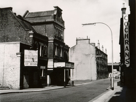 Black and white view along deserted street. A dilapidated theatre building to the left features a hoarding advertising the boy scouts of paisley gangshow of 1959. A raised railway line can be seen at the end of the street.