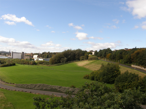 Colour view of open, grassy slope with unmarked roads at the top and bottom. Behind are some industrial buildings, set among trees, with a church spire and hills in the distance.