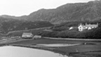 Black and white view showing a pond or partially flooded field, separated from another body of water by a built up road or causeway. Behind the field stands a small boathouse and a larger two-storey house, set among trees.